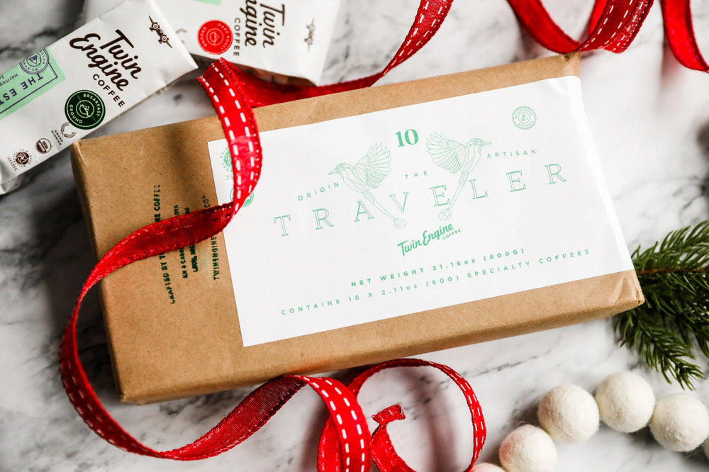 A kraft paper gift box with a white label reading 'The Traveler' and 'Twin Envelope', wrapped with a red ribbon, styled on a marble surface with coffee bags and holiday greenery.