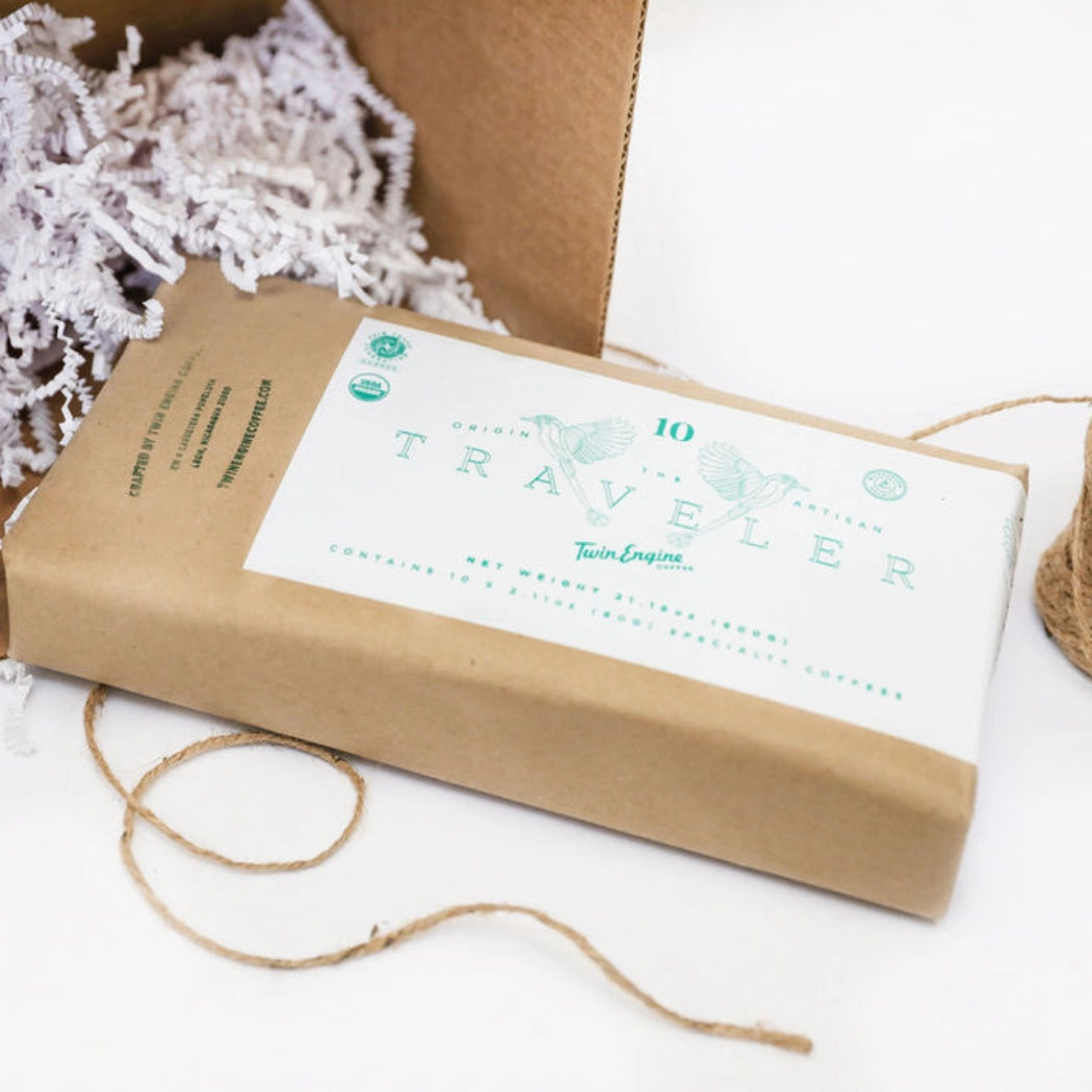 A kraft paper wrapped package labeled 'Traveler' with a white label and mint green text, placed on a white surface with jute twine and white filler material in a box.