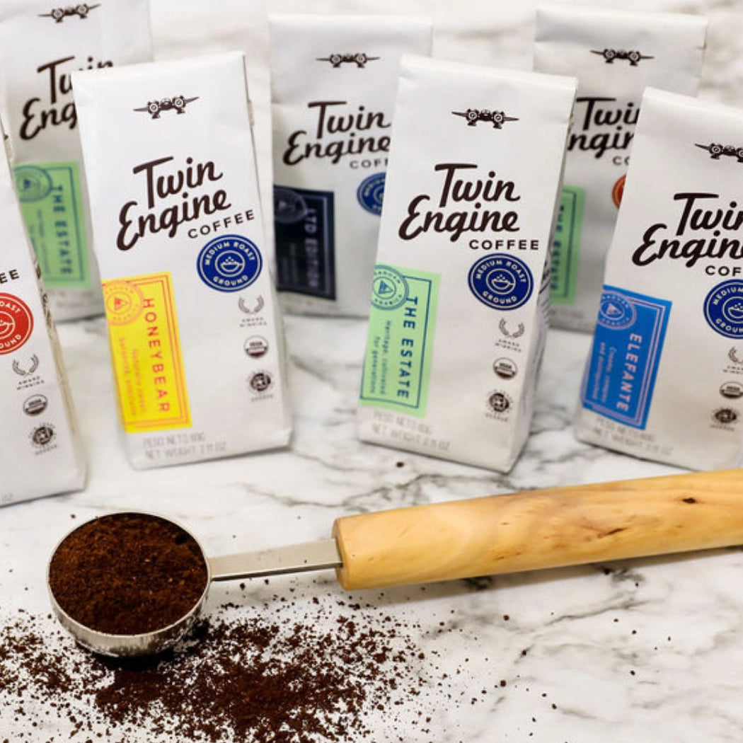 Several Twin Engine Coffee bag packages are arranged on a white marble surface with coffee grounds and a wooden-handled measuring scoop in the foreground. Visible bag colors include yellow, green, blue, and dark gray, while the packages themselves are white with black branding.