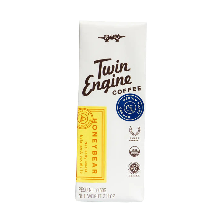 A single pack of Twin Engine Coffee Honeybear Medium Roast Ground Coffee, shown on a clean white background. The packaging is primarily white with dark brown text, a yellow vertical label, and a blue circular seal.
