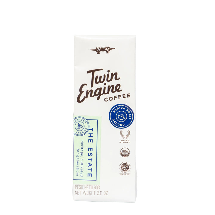 A package of Twin Engine Coffee 'The Estate' medium roast ground coffee, shown on a clean white background. The packaging is primarily white with brown lettering, a blue circular seal, and a light green vertical banner.