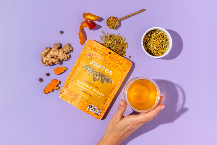 Jureea tea tumeric ginger packaging with a cup of tea, dried herbs, and a spoon on a purple background
