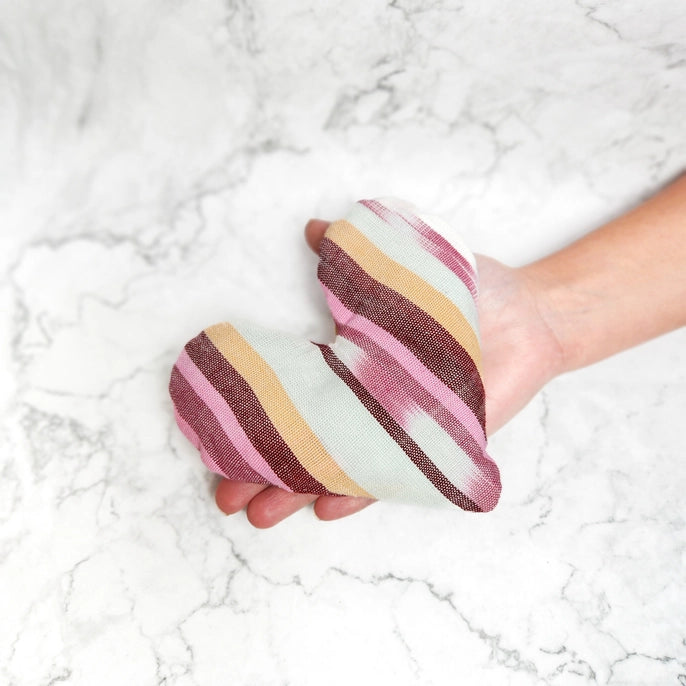 Hand holding a striped fabric heart shaped eye bag on a marble surface