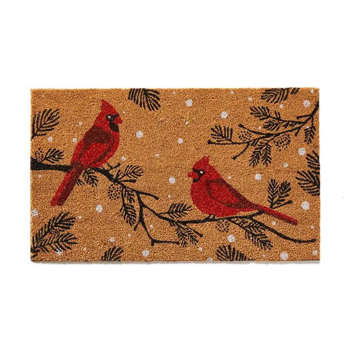 A rectangular coir doormat featuring two red cardinals perched on black pine branches with white snowflake details, shown against a plain white background.