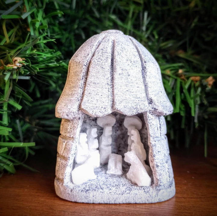 Small nativity scene figurine with a thatched roof carved from alabaster against a green foliage background