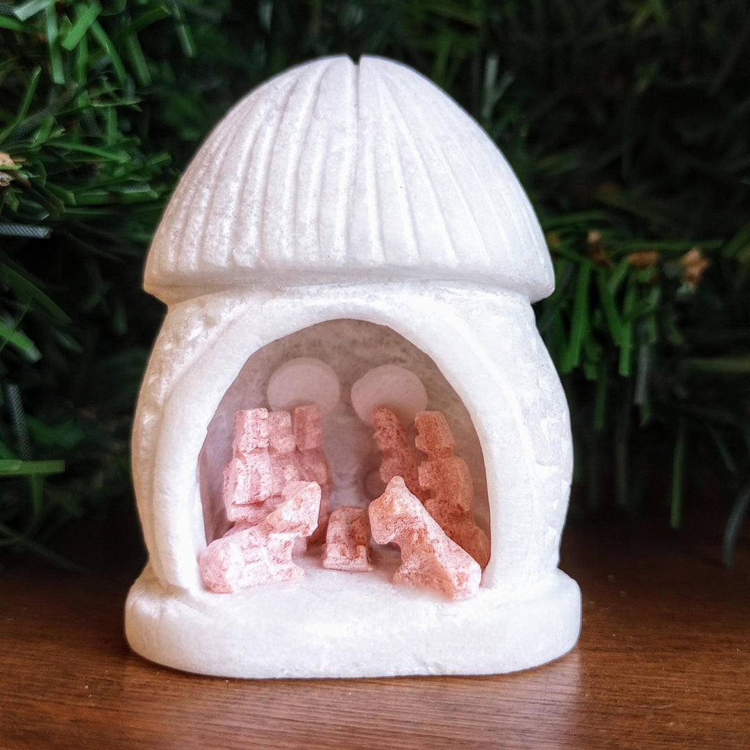 Small white nativity scene figurine with pink figures carved from alabaster on a wooden surface with greenery in the background