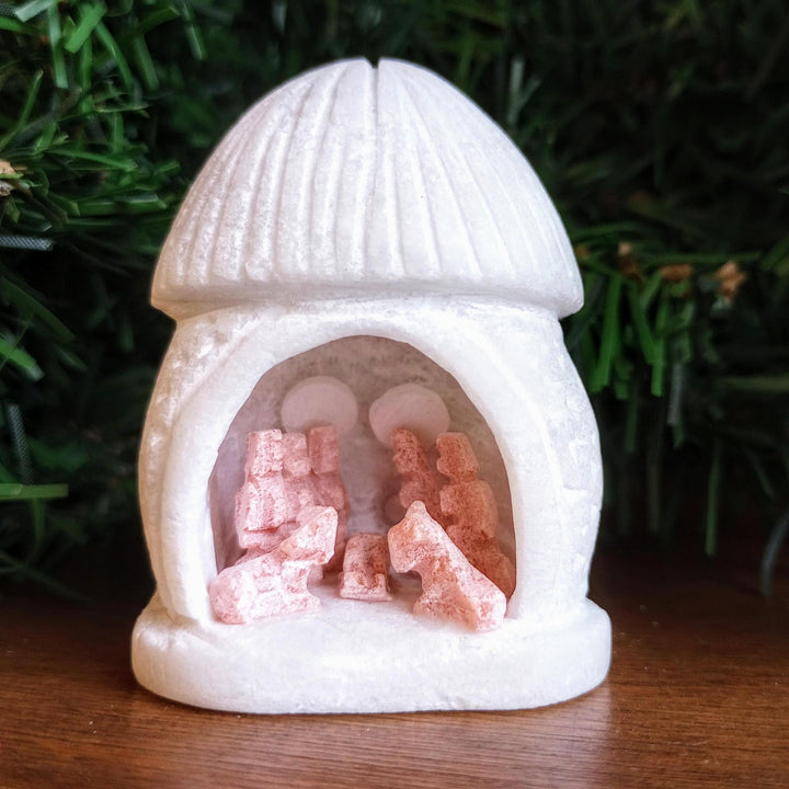 Small white nativity scene figurine with pink figures carved from alabaster on a wooden surface with greenery in the background