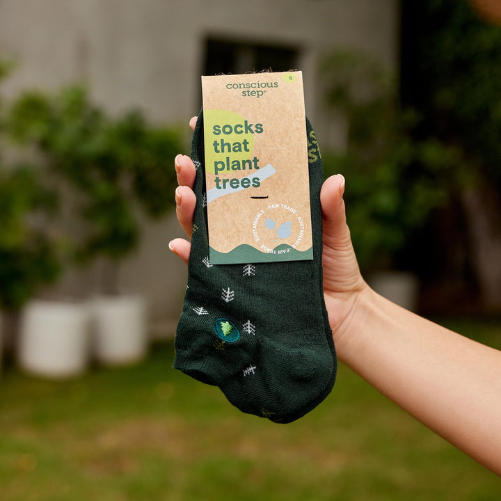 Hand holding a green sock with a 'conscious step' label, set against a blurred outdoor background.