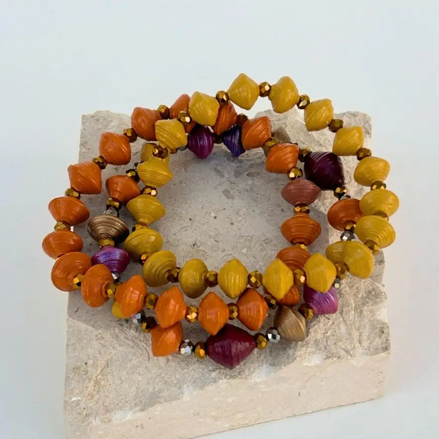 Three beaded stretch bracelets with orange, yellow, and purple shell-shaped beads and small metallic spacer beads, displayed on a light stone block against a white background.