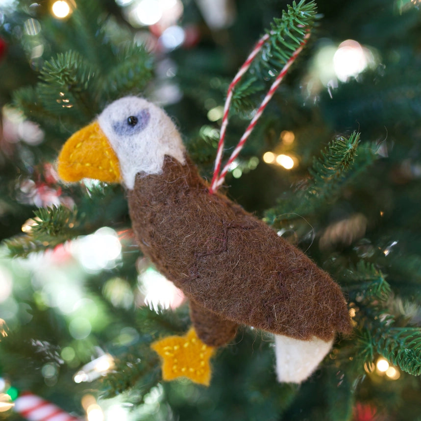 A felted wool bald eagle Christmas ornament hanging on a decorated Christmas tree with warm string lights in the background.