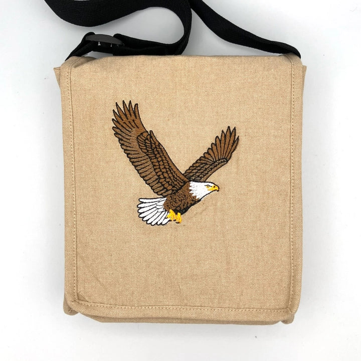 A beige canvas shoulder bag with a black adjustable strap, featuring an embroidered bald eagle design on the front pocket.