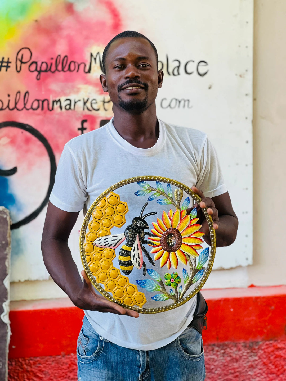 Artist Achelet holding a round hand-painted decorative metal wall featuring a yellow sunflower, a bumblebee, and a honeycomb pattern, set against a graffiti-covered wall.