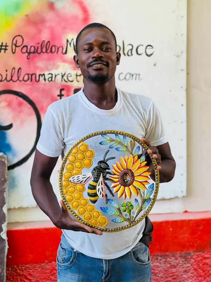 Artist Achelet holding a round hand-painted decorative metal wall featuring a yellow sunflower, a bumblebee, and a honeycomb pattern, set against a graffiti-covered wall.