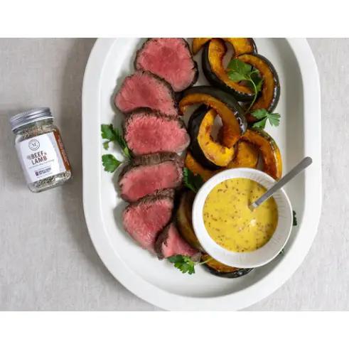 Plated dish of sliced beef with roasted vegetables and a bowl of yellow sauce, accompanied by a jar of Martha Stewart beef and lamb seasoning on a light gray background.