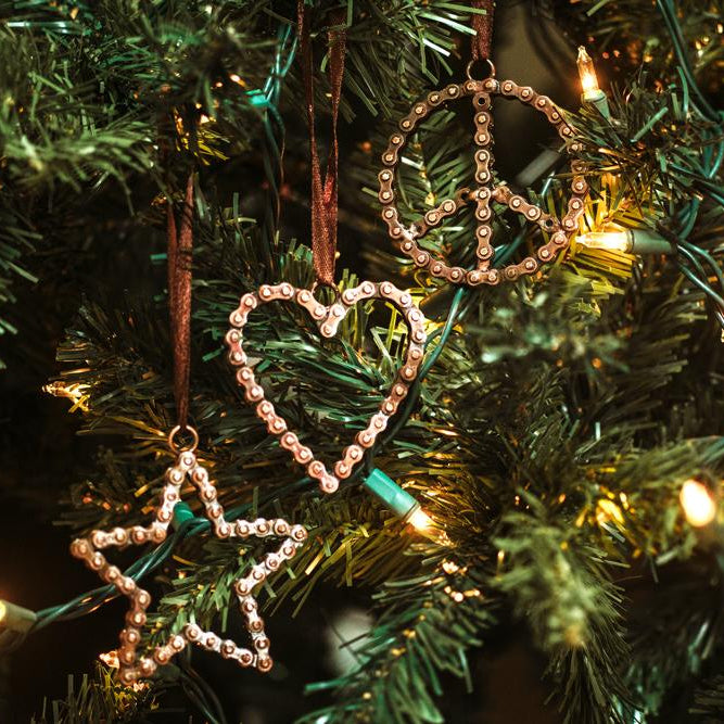 Three Christmas tree ornaments in the shapes of a peace sign, a heart, and a star, hanging on a green Christmas tree with warm white string lights.