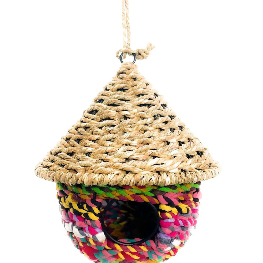 A colorful bird bungalow made from seagrass and sari fabric, with a thatched roof and a round opening for birds to enter.