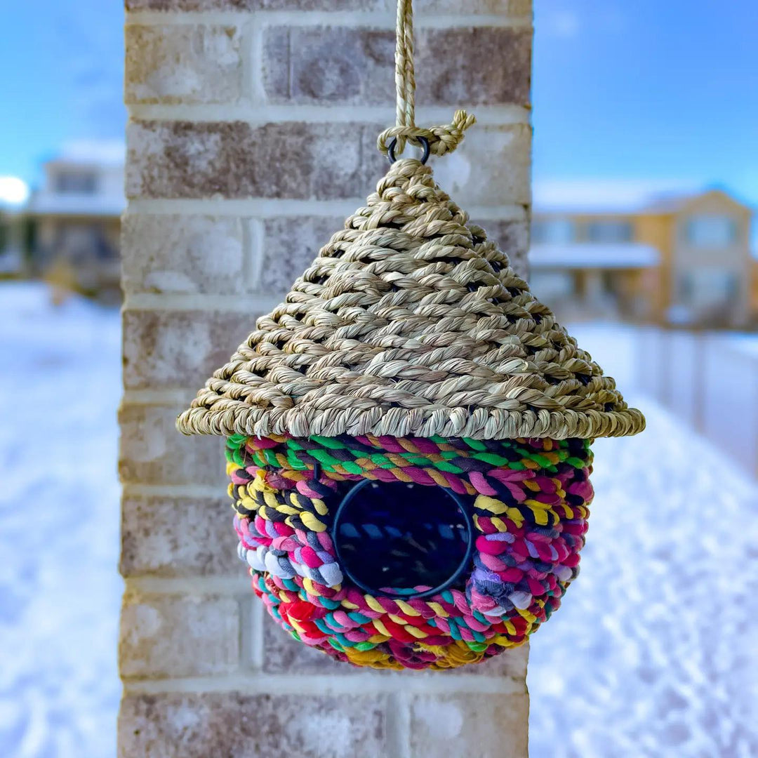 A colorful hanging bird bungalow made from seagrass and sari fabric, with a thatched roof and a round opening for birds to enter.