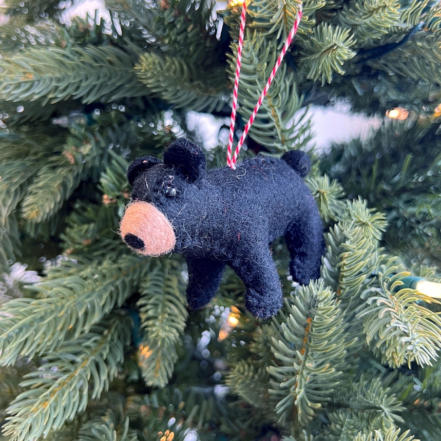 A small black felt bear ornament with a tan snout hangs from a red-and-white striped string on a green Christmas tree.