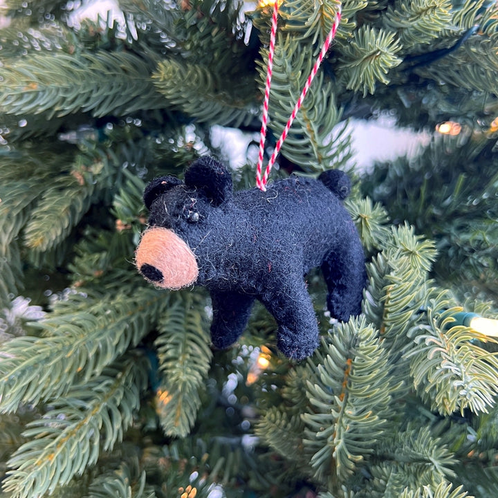 A small black felt bear ornament with a tan snout hangs from a red-and-white striped string on a green Christmas tree.