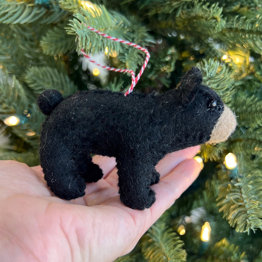 A small black felt dog Christmas ornament with a beige snout and a red-and-white hanging string, held in a person's hand in front of a lit Christmas tree.
