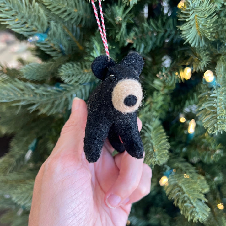A hand holds a small black felt bear ornament with a beige snout and nose, hanging from a red-and-white striped string in front of a green Christmas tree with warm lights.