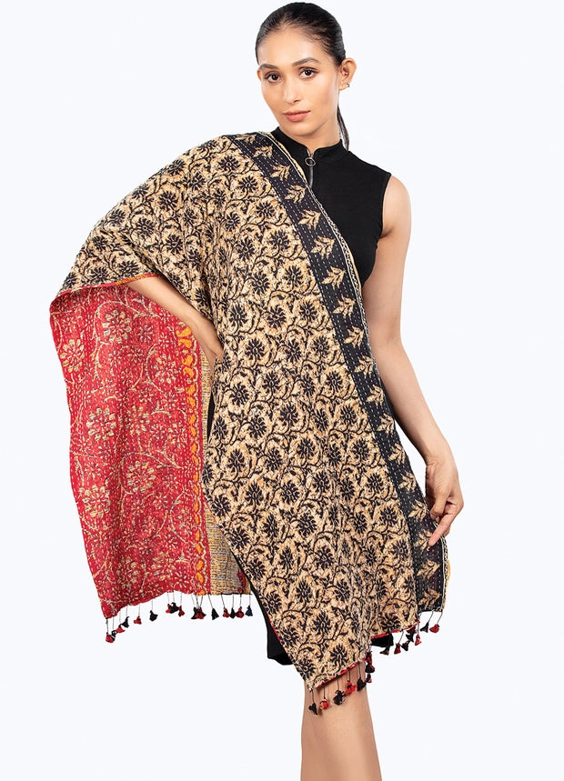 A woman models a beige and black floral-patterned shawl with a red inner lining and black tassels, worn over a black sleeveless top against a light gray background.