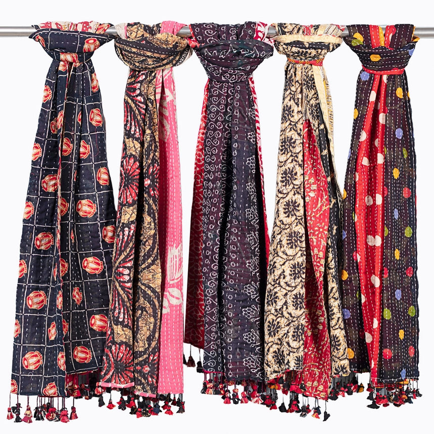 Five colorful patterned scarves hanging from a metal rod against a white background, each with tassels at the bottom.