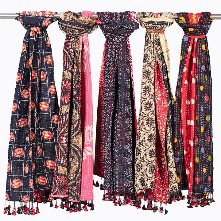 Five colorful patterned scarves hanging from a metal rod against a white background, each with tassels at the bottom.
