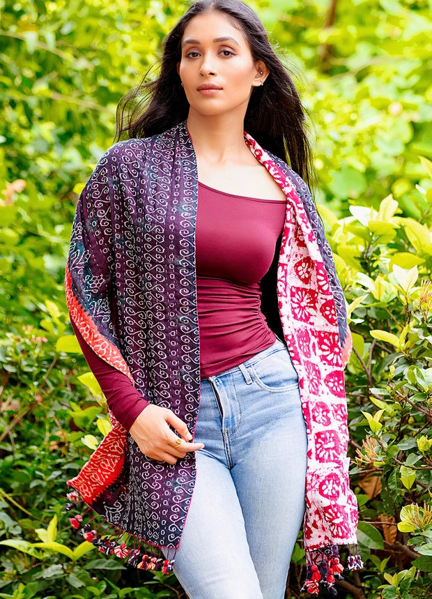 A woman wearing a dark purple patterned shawl with red and white accents over a burgundy top and light blue jeans, standing outdoors in front of a green garden background.