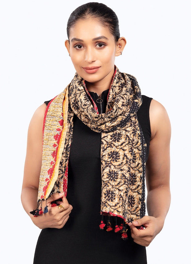 A woman modeling a patterned scarf with black floral motifs on a beige background, featuring red and yellow borders and tassels, paired with a black sleeveless top against a white studio background.