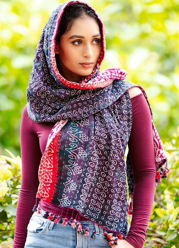 A woman outdoors wearing a patterned multicolor headscarf with tassel trim over a dark purple long-sleeve top and light blue jeans, with a blurred green foliage background.