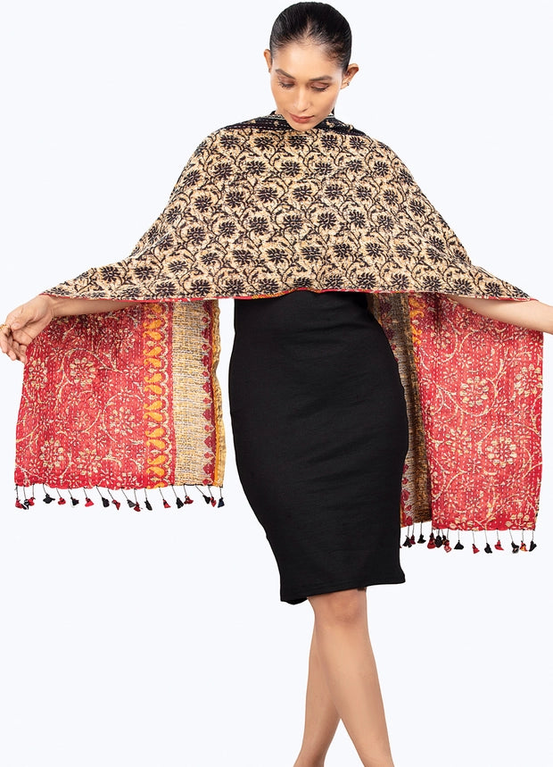 A woman modeling a large patterned shawl draped over her shoulders, with the black skirt and arms visible. The shawl has a beige and black floral pattern on one side and a red and gold patterned border with tassels on the other, set against a plain white background.