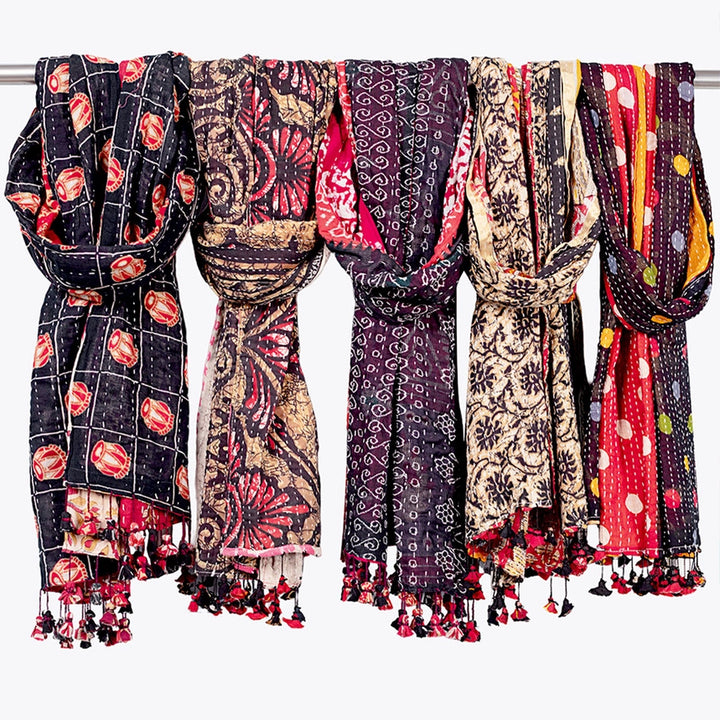 Five colorful patterned scarves with tassels hanging from a metal rod against a plain white background.