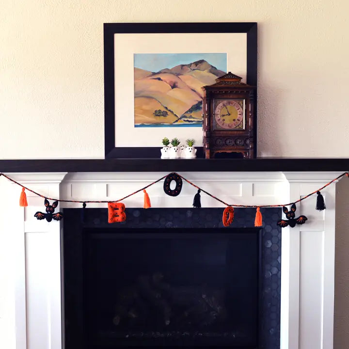 A framed landscape painting of rolling hills hangs on a beige wall above a white fireplace mantel decorated with a black mantel shelf, a dark wood clock, small potted plants, and a Halloween garland with orange letters and black tassels.