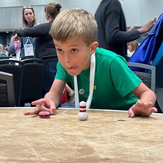 Young boy in a green shirt playing with small bowling pins on a table, with people in the background.