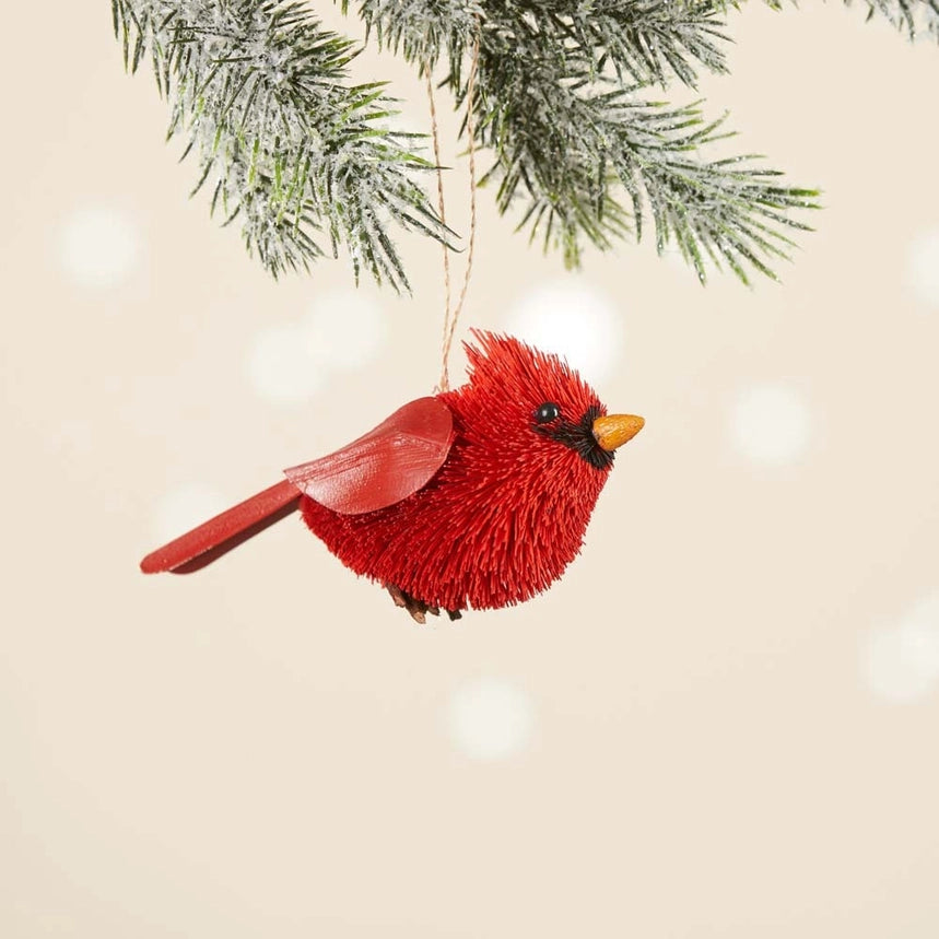 A red cardinal bird Christmas ornament with a textured, buri red body, a small yellow beak, and black eye details, hanging from a snowy green pine branch against a warm beige background with soft bokeh lights.