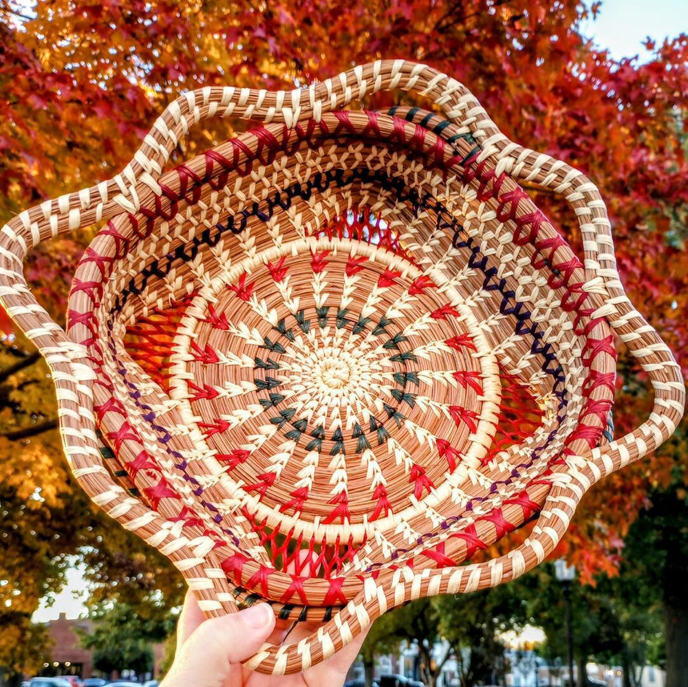 Handwoven basket with pink and red decorations with a spiral pattern with fall trees in the background.