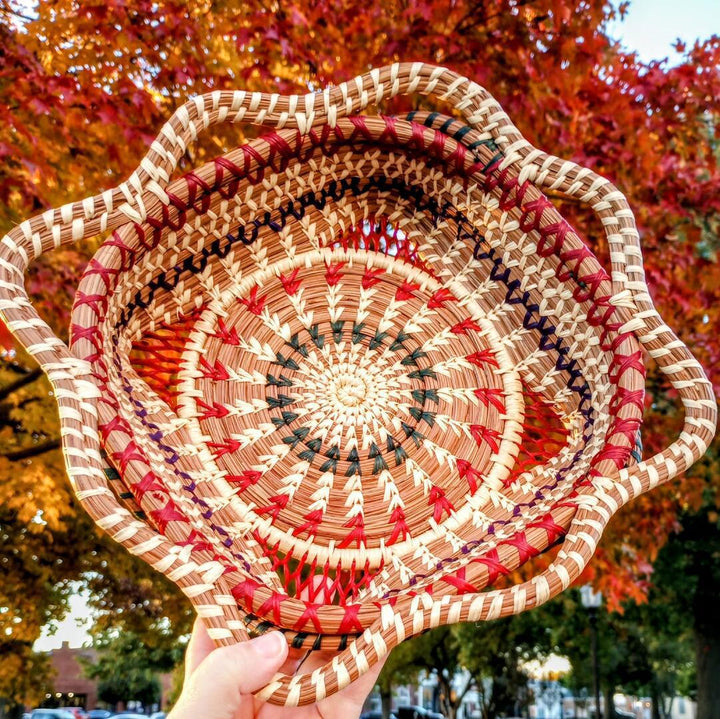 Handwoven basket with pink and red decorations with a spiral pattern with fall trees in the background.
