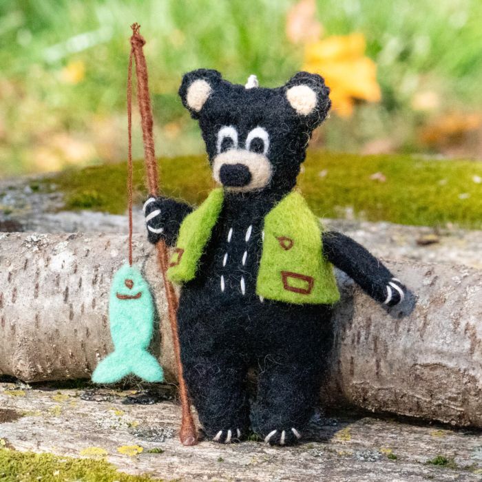 A felted bear in a green vest holding a fishing rod and fish on a natural background