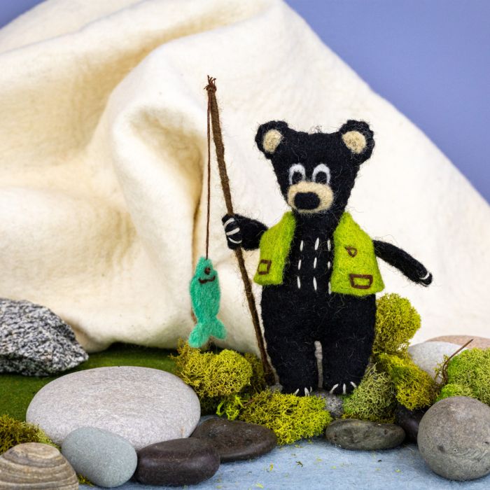 Black bear felted ornament with fishing rod and green vest on a natural setting with stones and moss.