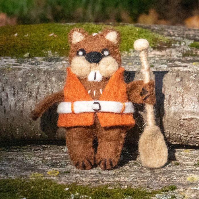 Felted animal figurine of a beaver in an orange jacket with a paddle, standing on a wooden surface with moss.
