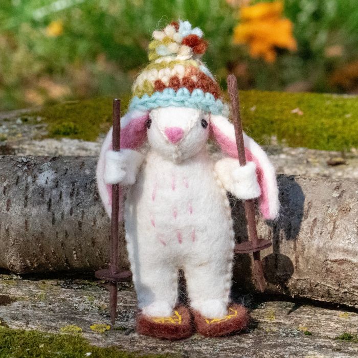 felted animal resembling a rabbit wearing a colorful hat and holding skis, standing on a snow shoes with a natural background.