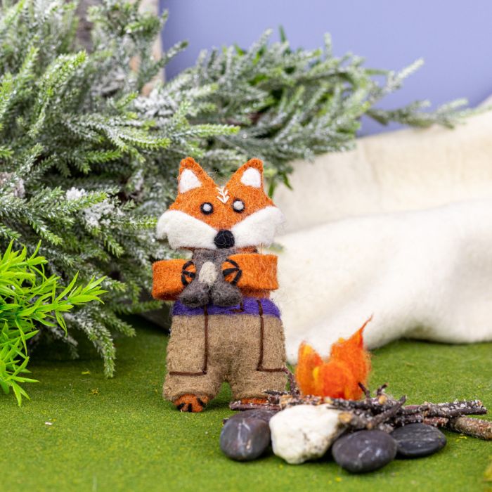 Felt fox ornament holding binoculars by a campfire with evergreen trees in the background