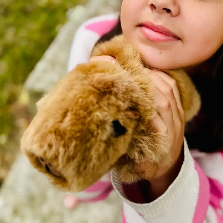 A close-up lifestyle shot of a person holding a brown plush capybara toy against an outdoor blurred background.