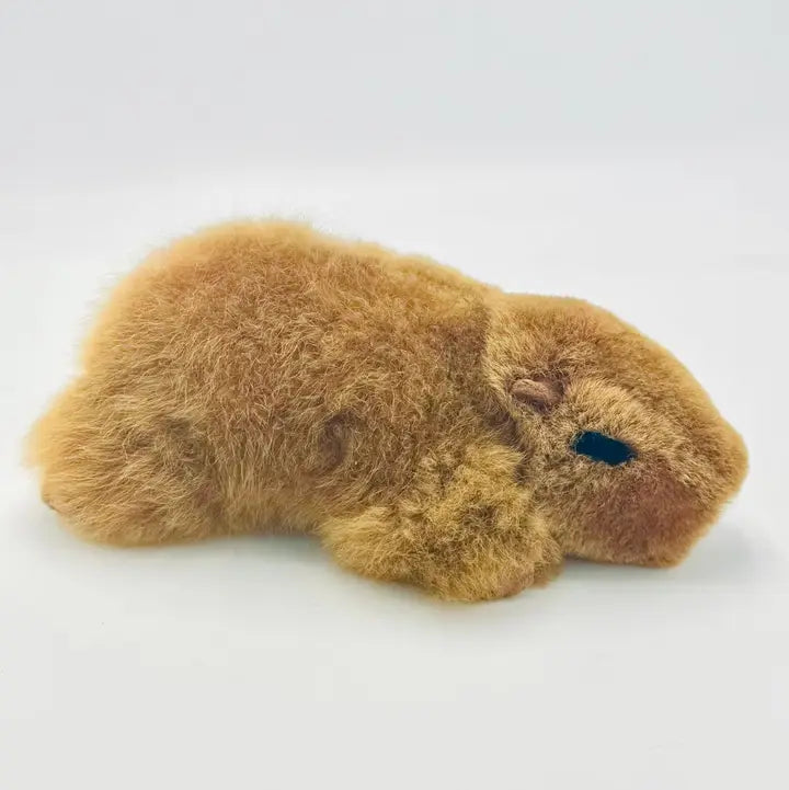 A small plush capybara toy with fluffy tan-brown fur and a small dark blue embroidered eye, photographed on a plain light gray background.