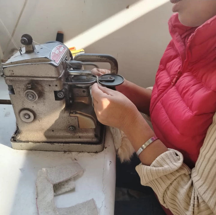 A close-up of a person wearing a red puffer vest and beige ribbed sweater operating a vintage, worn sewing machine with a metal cover open, set against a plain indoor background.