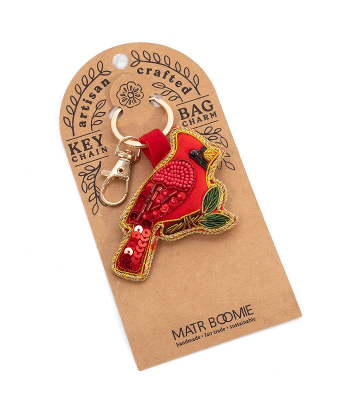 A red cardinal bird keychain charm with gold sequins and a red fabric hood, attached to a brown kraft paper display card that reads 'MATR BODMIE'.