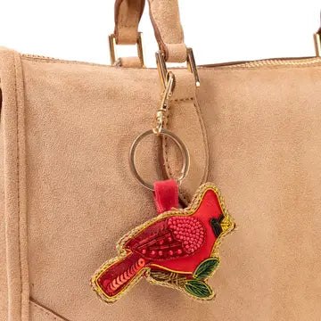 A close-up of a tan suede handbag with a red cardinal-shaped embroidered keychain charm hanging from a gold-tone ring.