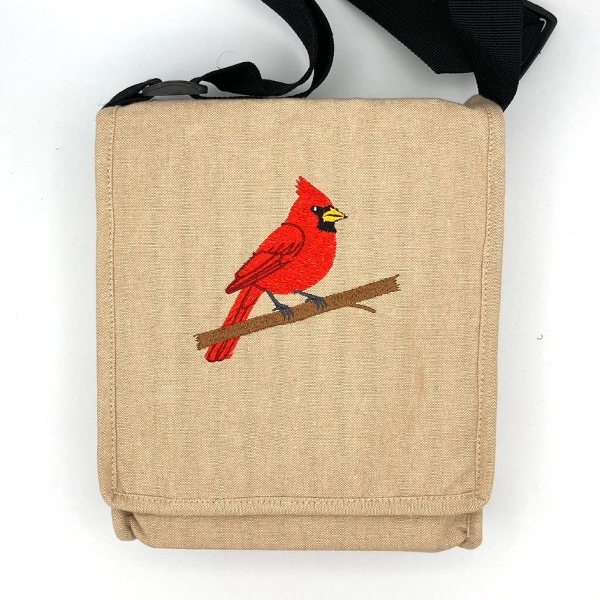 A beige canvas shoulder bag with a black strap, featuring an embroidered red cardinal bird on a brown branch on the front pocket, set against a plain white background.
