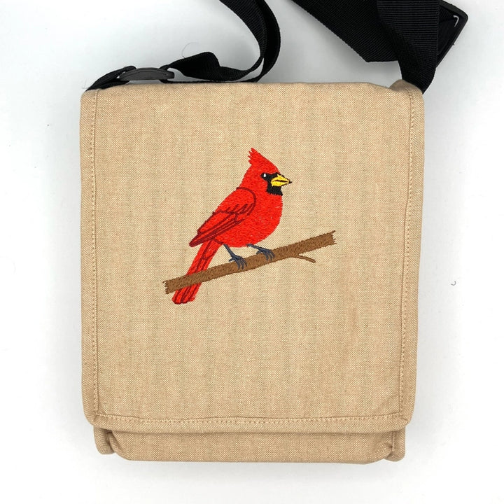 A beige canvas shoulder bag with a black strap, featuring an embroidered red cardinal bird on a brown branch on the front pocket, set against a plain white background.